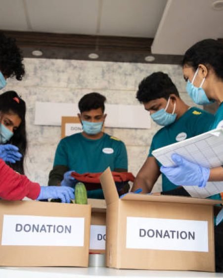 Low angle view, group of volunteers busy working by arranging vegetables and clothes on donation boxes and noting down during coronavirus covid-19 pandemic lockdown for needy people